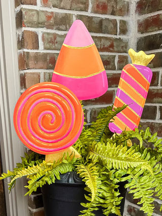 A decorative autumn arrangement featuring colorful candy-shaped and pumpkin-shaped yard decorations in pink, orange, and yellow, styled in a black urn planter with green ferns on a brick porch next to a wooden door.